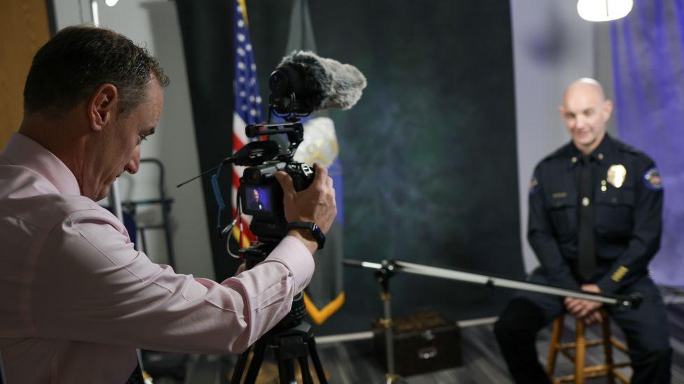 An Aurora police officer in uniform sits on a stool in a studio as a man in a pastel work shirt adjusts the camera filming him, an American flag visible in the background