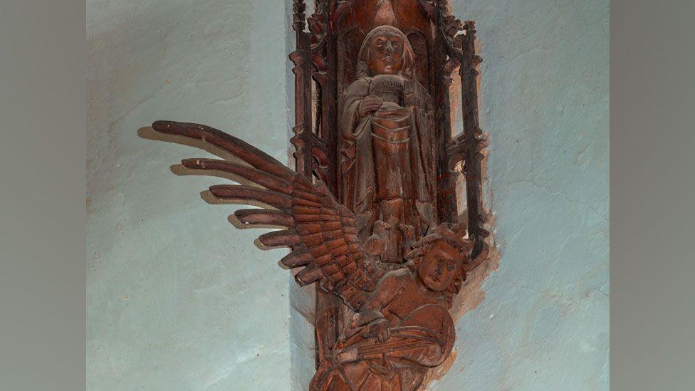 A wooden carving of an Anglo-Saxon saint. She is standing against a wall and at her feet are birds. Beneath her feet is an angel holding a stringed instrument with one wing extended left