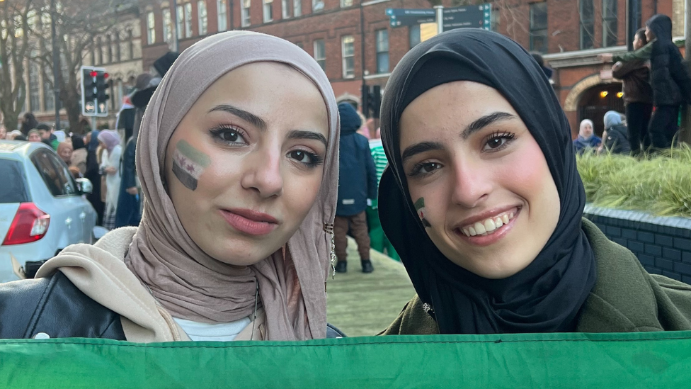 Two girls smiling, girl on left is wearing a pink headscarf, black leather jacket and cream hoody, girl on right (Renad) is wearing a black headscarf and dark green jacket, both girls have syrian flags painted on their faces, group celebrating in the background