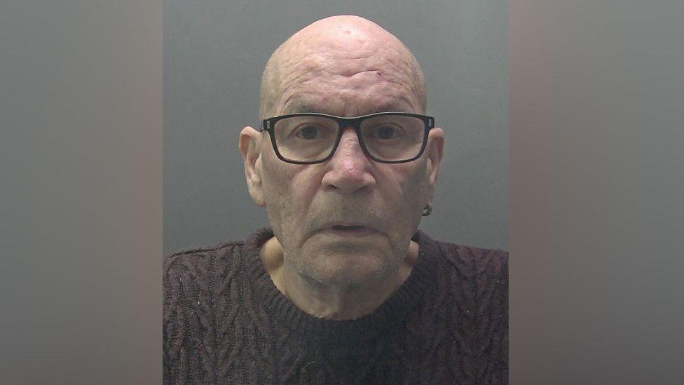 Custody photograph of David Newton, who is clean shaven and bald, looking into the camera, wearing dark glasses, a ring-shaped earing and purple jumper