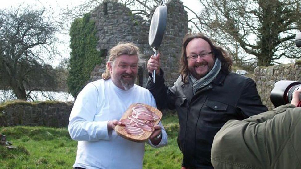 Si and Dave stand posing for the camera. Si is dressed in a white top and is smiling. He is holding a tray of bacon. Dave stands next to him and is wearing a leather jacket. He has a big grin on his face and is holding a frying pan above his head.