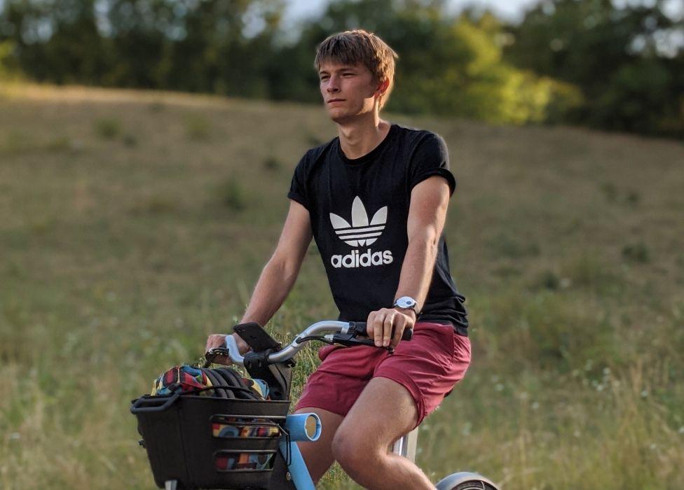 A young man wearing an adidas top cycles on a blue bike, with a field in the background