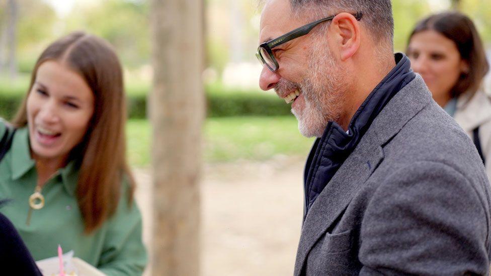 Lidia is in a pea-green top with a gold zip, Cristina in a white jacket, and Felix in a blue jacket - all stand laughing with Victoria (unseen) in a park