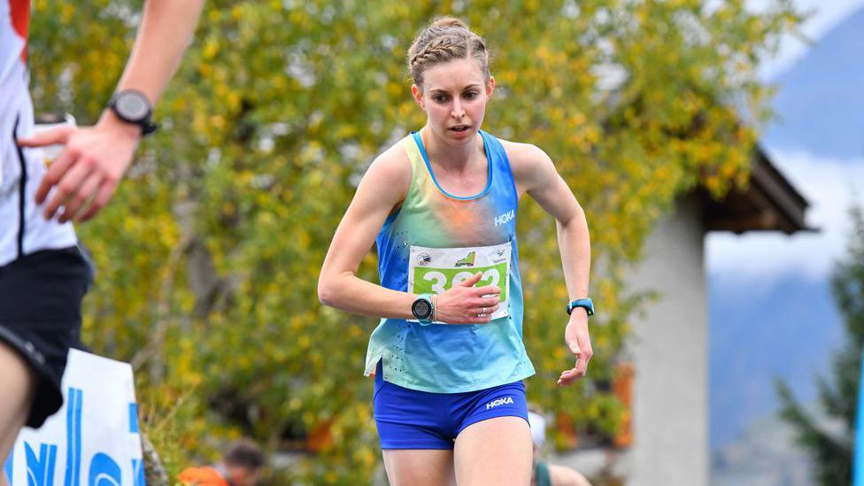 A close-up of a runner in a multi-coloured top looking determined to keep up a good tempo with a tree and house in the background