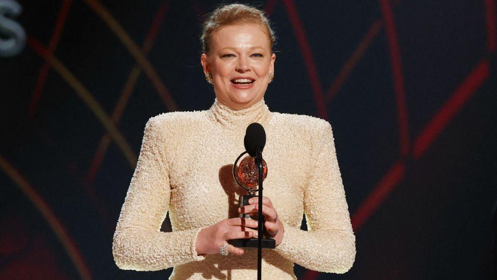 Sarah Snook accepts the Best Performance by an Actress in a Leading Role in a Play award for The Picture of Dorian Gray at the the 78th Annual Tony Awards in New York. she is wearing a cream high-neck dress with long sleeves.