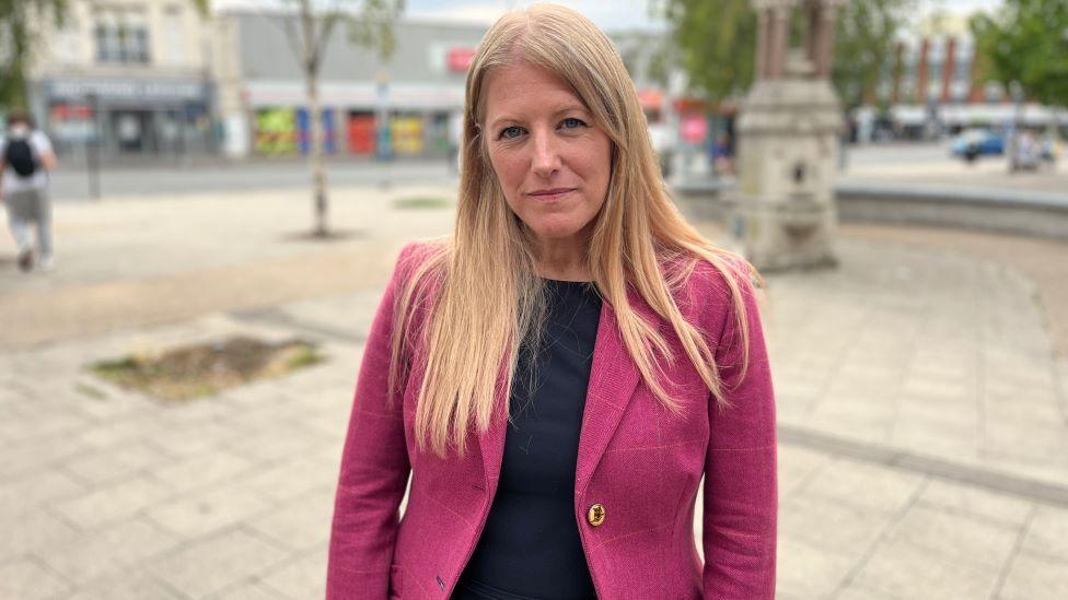 A woman with long blonde hair, in a pink blazer, stands in an empty town square, looking at the camera.