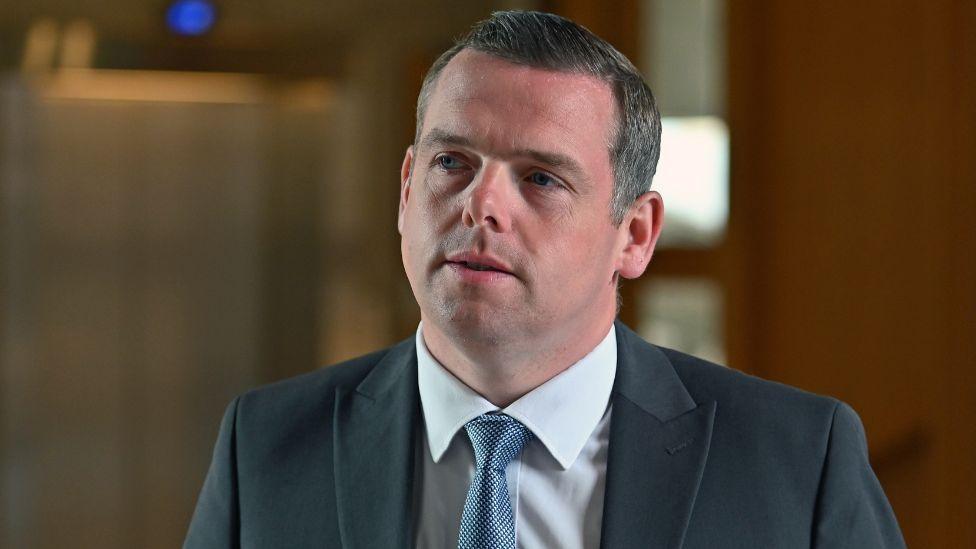 Douglas Ross, with dark hair and wearing a dark suit, in a medium close-up shot in the Scottish Parliament 