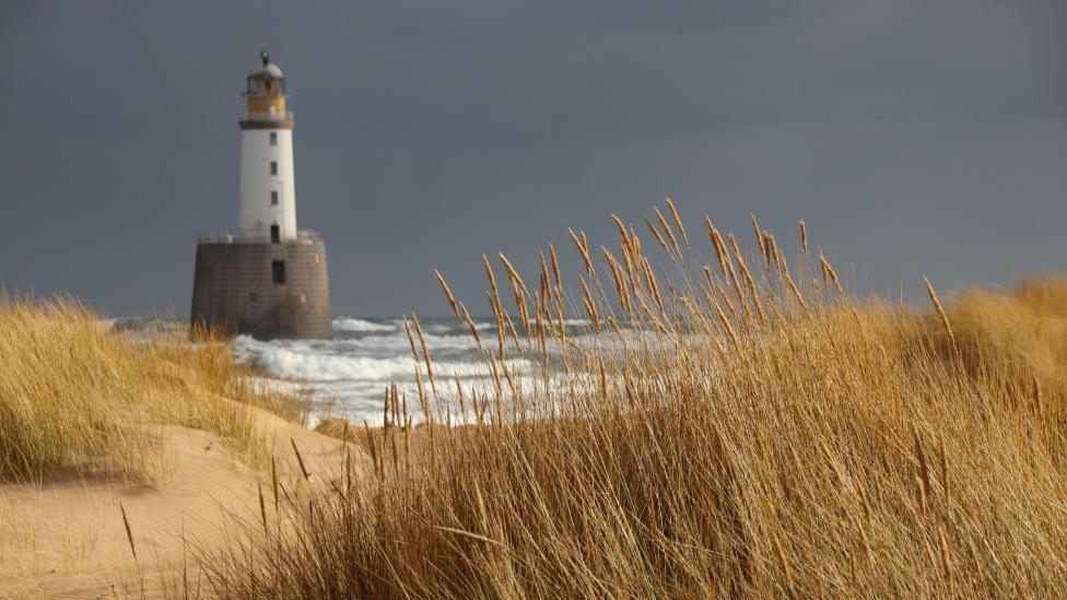 White lighthouse on a grey base, in sea surrounded by waves, taken from grassy beach location.