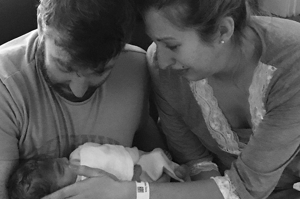 Black and white photo of Dan and Fiona cradling their newborn baby. Dan holds her in his arms and is looking down at her, while Fiona strokes her face. A hospital band is visible on her wrist.
