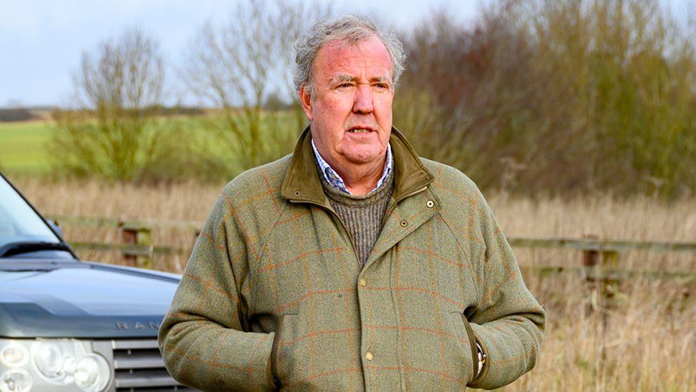 Jeremy Clarkson standing in a field in front of a Range Rover car looking into the distance wearing a green tweed coat