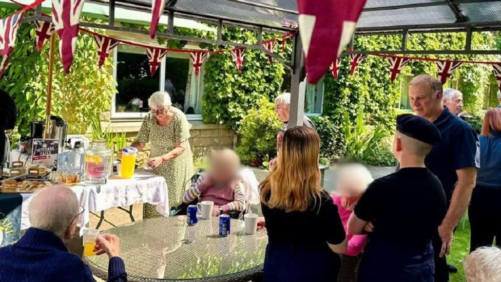 People of varying ages sit at tables in a garden that has bunting up. A woman wearing a green summer dress pours a drink and other people sit drinking cups of tea and other drinks.