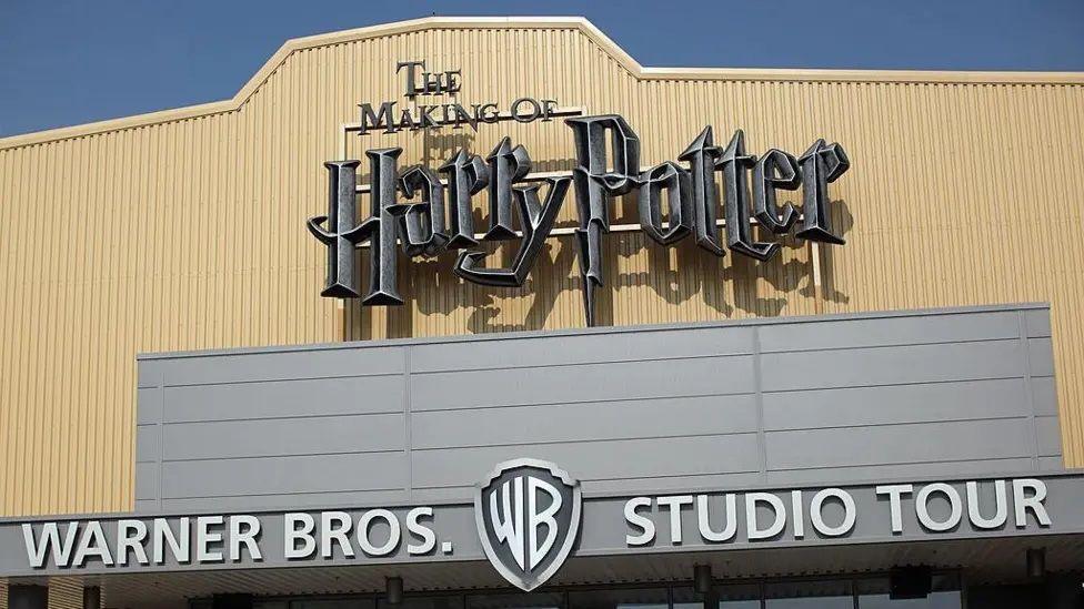 The exterior of the Warner Bros Studio Tour.
A large silver sign says "The Making of Harry Potter" in the lettering used on the movies, and below that in a normal font "Warner Bros. Studio Tour" with the WB logo in the middle. IT is the top of the building in cream with a blue sky behind.