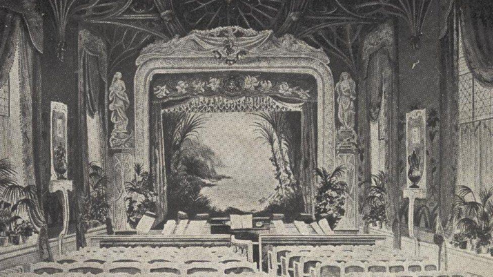 Black and white photo of the Gaiety Theatre at Plas Newydd, Anglesey. There are rows of seats in lines of five and six in front of an orchestra pit and a proscenium arch stage with curtains and a painted front curtain showing an island and seascape. The room has vaulted arches on the ceiling and lots of palm plants dotted around the edges.