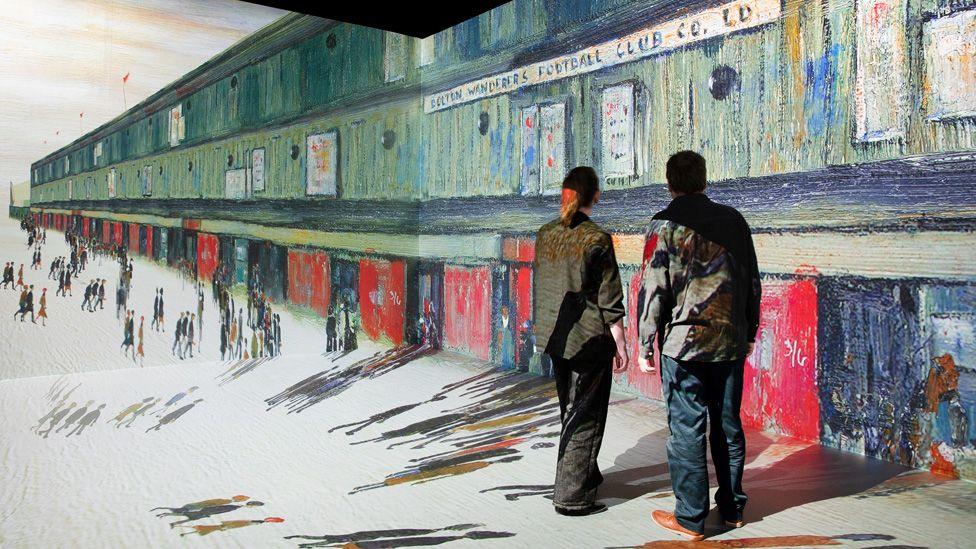 Two people standing in a room with LS Lowry's Going to the Match projected onto the walls and floor
