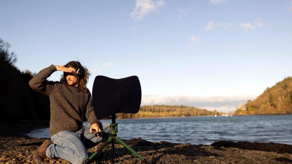 A long-brown haired man looks to the sky. He is surrounded by water and mountains. He holds a large black sound recorder, that resembles an umbrella. He wears black headphones, a black t-shirt and a blue jumper.