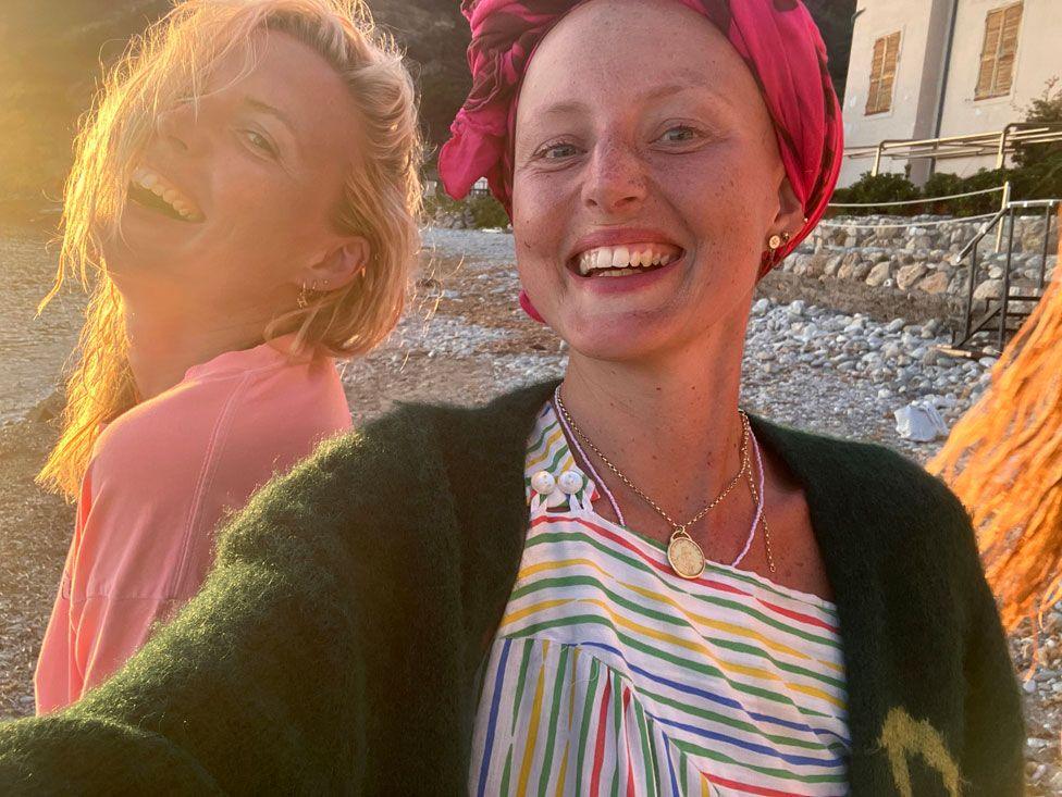 Kris and Maren are taking a selfie. Kris is wearing a rainbow striped top with a green cardigan. She has a pink scarf wrapped around her head. Maren is wearing a pink jumper. They are smiling and look joyful.