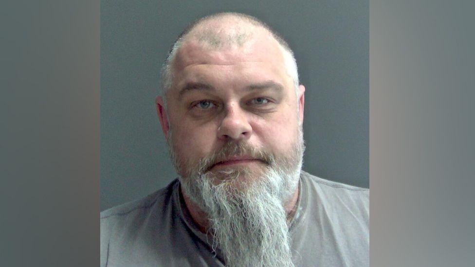 Colin Day is looking at the camera in a police custody shot. He has a balding head, a long grey beard and a light brown moustache. He is wearing a grey T-shirt.