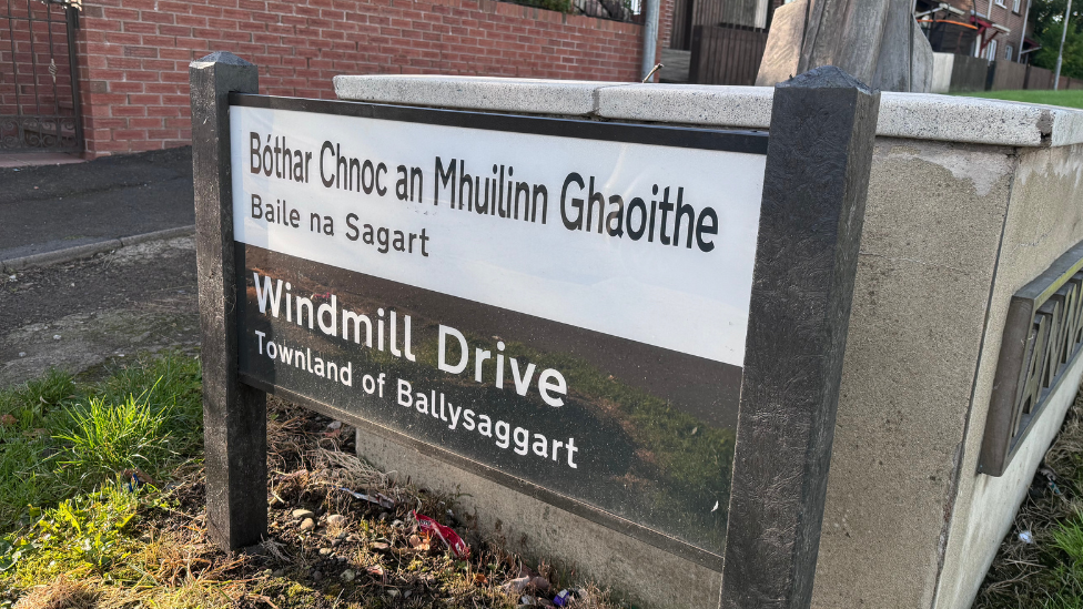 A Windmill Drive street sign - at the top Windmill Drive is written in Irish in black font against a white background. On the bottom in a black background in white font Windmill Drive is written in English. The sign is in front of a grey wall.