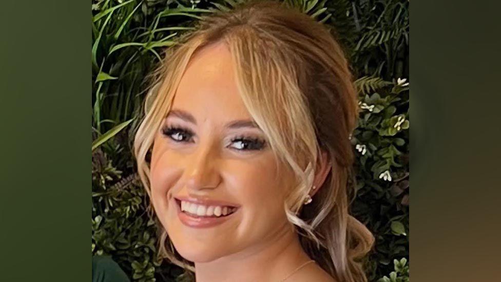 A woman, with blonde hair wearing makeup and fake eyelashes. She is smiling and standing in front of some green leaves.