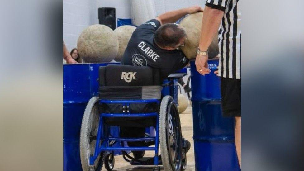 Mr Clarke in his wheelchair and his back to the camera as he lifts weighted balls. He is wearing a navy blue t shirt with his surname Clarke written on it in white writing. There is what looks like an umpire beside him wearing a black and white striped t shirt and black shorts.
