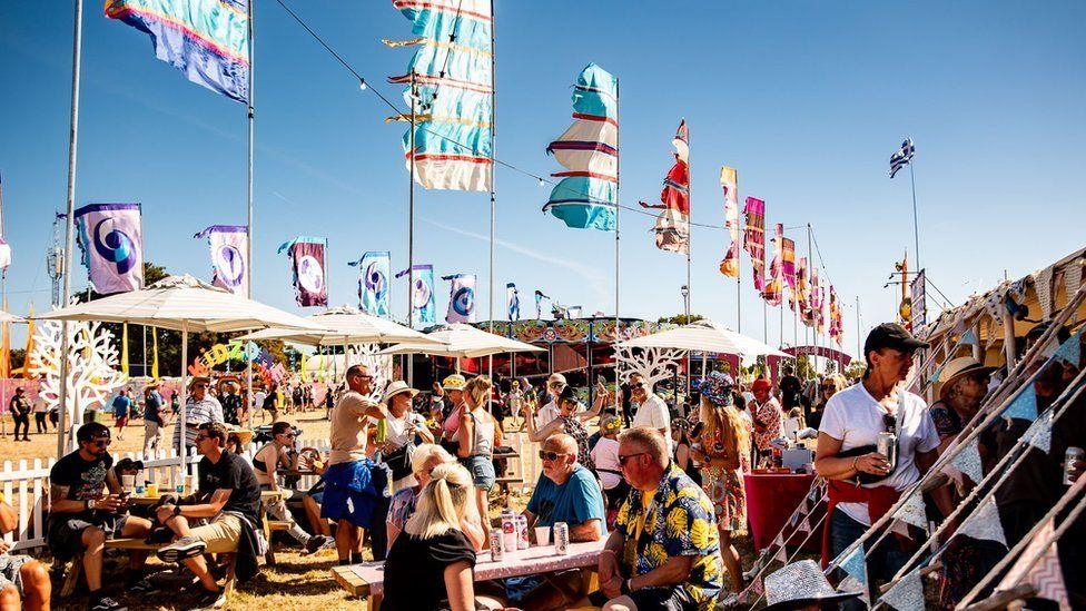 Isle of Wight festivalgoers sitting at tables with tents and colourful flags blowing in the wind