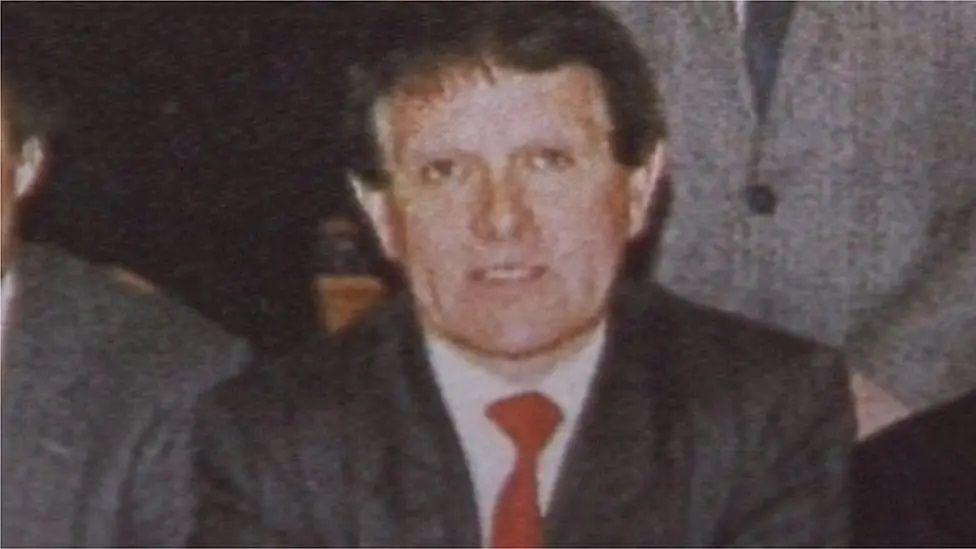 A man with short brown hair wearing a grey blazer, white shirt and red tie