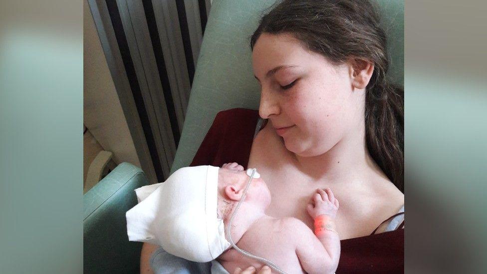 Freya with her brother Harry after he was born. Harry has a tube and a white stocking hat and is layign on Freya's chest as she sits up.