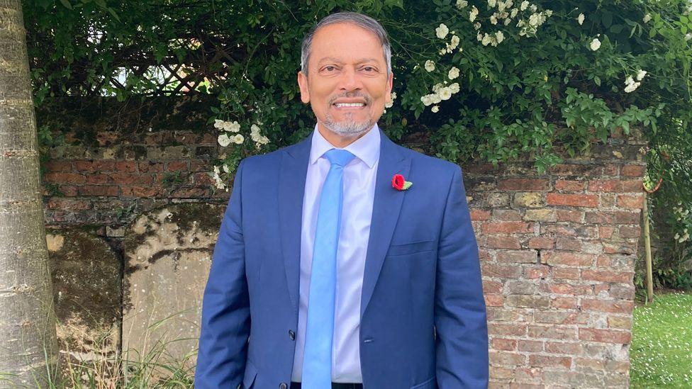 Chan Abraham wearing a navy suit and light blue tie standing next to a tree