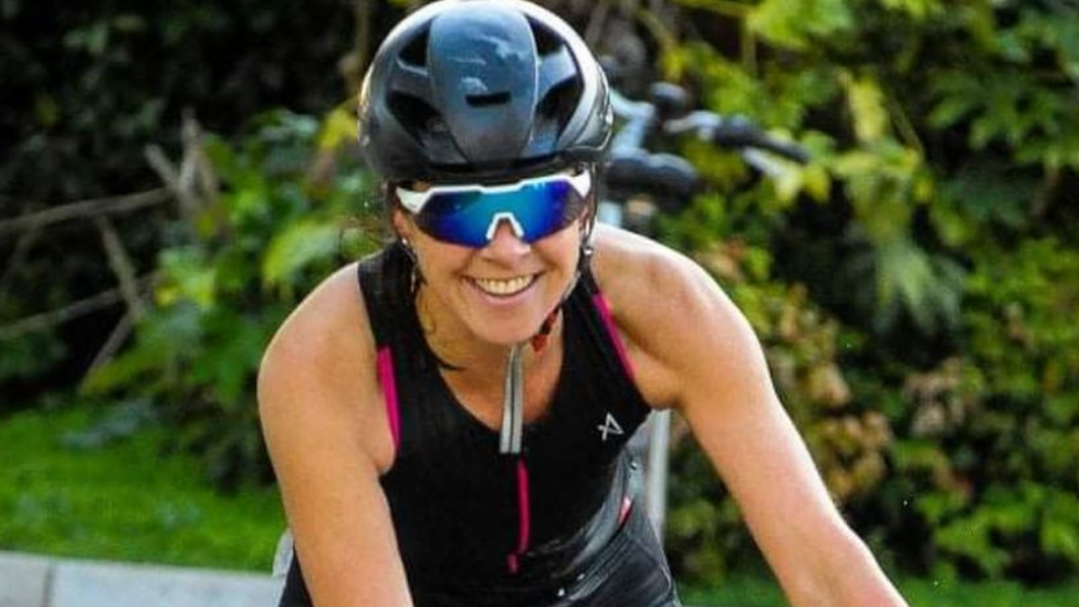 Rebecca Comins cycling and grinning with a bike helmet and sunglasses on 