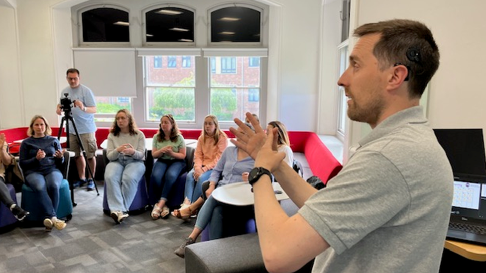 Man stands in class room as students watch on seated in a circle