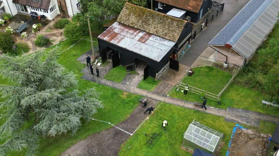 Aerial view of Stocking Farm as police search for body of Muriel McKay in July 2024. Foreground shows a tall fir tree surrounded by grass. Behind it a barn building with black walls and a metal roof, and a simlar building behind that. Right foreground there is a greenhouse and next to that a dug up patch of soil. Police officers are seen from above wearing black clothing