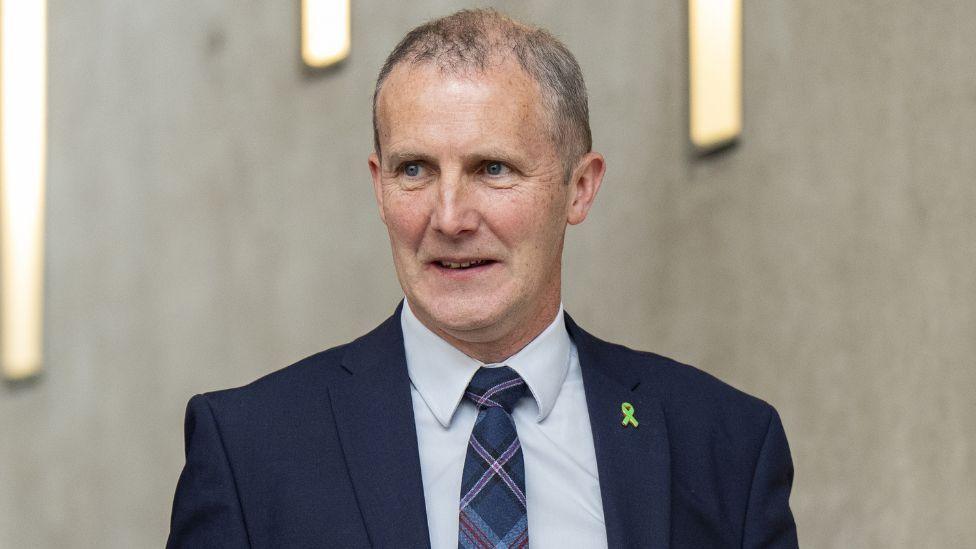 Michael Matheson, with thinning grey hair and wearing a dark blue suit, light blue shirt and blue tartan tie, looks to his right while walking in the Scottish Parliament 