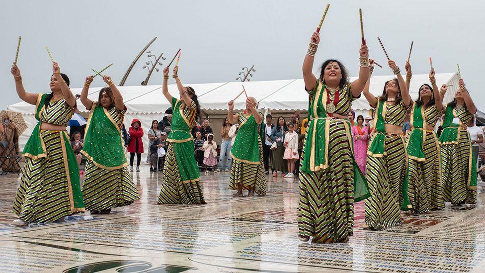 Indian dancers in two lines, in long, green dresses dancing on the comedy carpet in Blackpool, holding sticks above their heads.