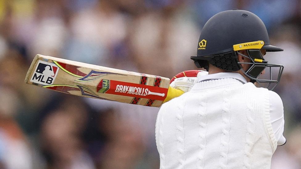 England cricketer Harry Brook stands with his back to the camera, bat over his shoulder, awaiting a bowl. He's wearing a cricket helmet, cricket whites and padded cricket gloves. The bat is a standard design with a Gray-Nicolls logo down the middle, and two chequerboard stripes around the top. There's also a square sticker at the end of the bat, displaying the MLB (Major League Baseball) logo. It's got the letters MLB in blue block capitals along the bottom half. The top half has the white silhouette of a baseball player, his bat cocked as a ball approaches.