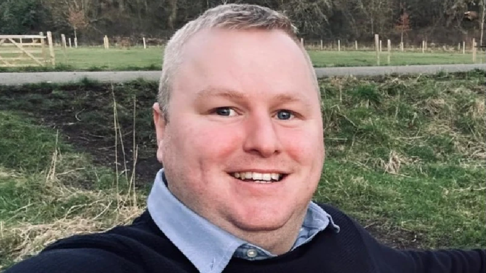 Darren Brown who has short grey hair, is wearing a light blue shirt and navy jumper and is smiling at the camera 