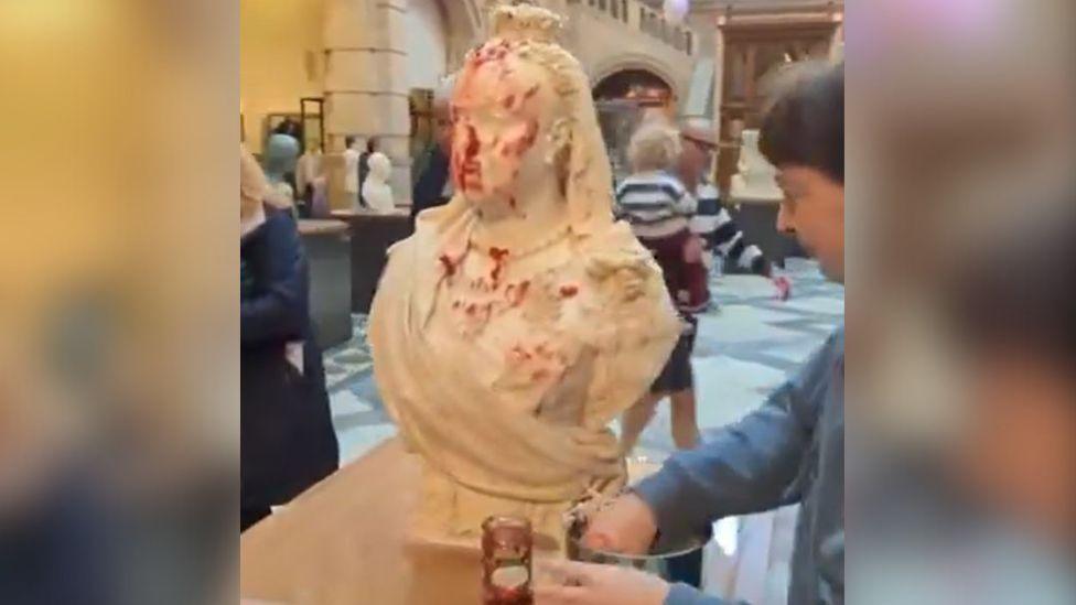 A woman with dark hair and a grey jumper spreads jam and porridge on a bust of Queen Victoria in a museum 