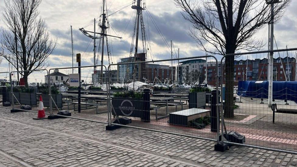 A cobbled street with a tall metal barrier fencing off trees and a pedestrianised walkway. In the distance are boat masks and behind them is a row of tall flats and commercial buildings.