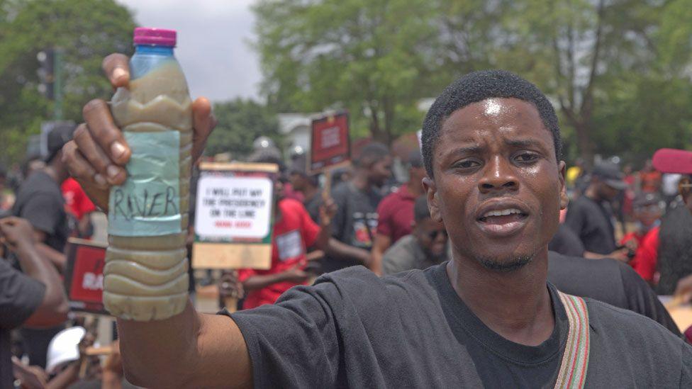 A protester wearing a black T-shirt holds up a bottle of dirty water. Other people can be seen behind him