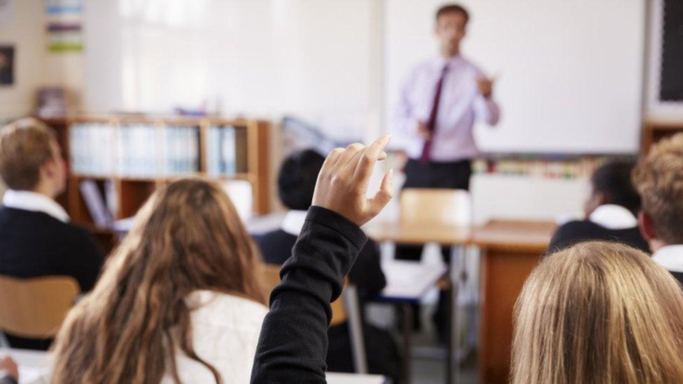 Teacher standing at front of classroom with child's hand raised in foreground