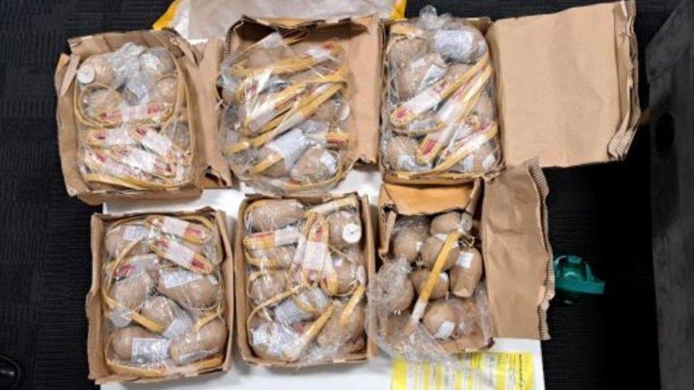Dozens of lightbulb-shaped parcels wrapped in brown paper sit in six cardboard boxes. There is plastic wrapped tightly around the small items. 