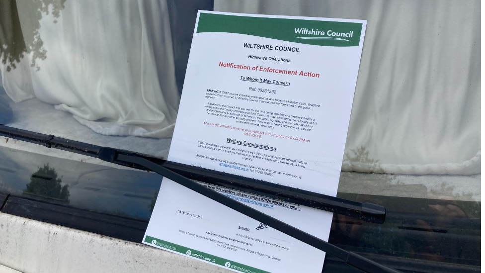 A notice tucked under a windshield viper. It is a letter from the Wiltshire Council, notifying the vehicle owner of 'Enforcement Action'.