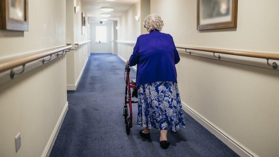 An elderly woman walks away from the camera along a corridor in a care home. Wearing a blue floral skirt and blue cardigan, she is using a walking frame. The carpet is blue and the walls are white with hand rails along the length of them.