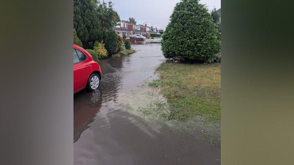 Flood water up someone's drive, surrounding a red car. In the distance there is flood water along a residential road