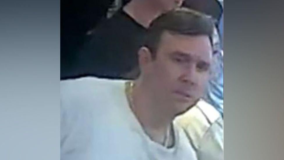 A grainy image shows a white man with short dark hair. He is wearing a gold necklace and a white t-shirt