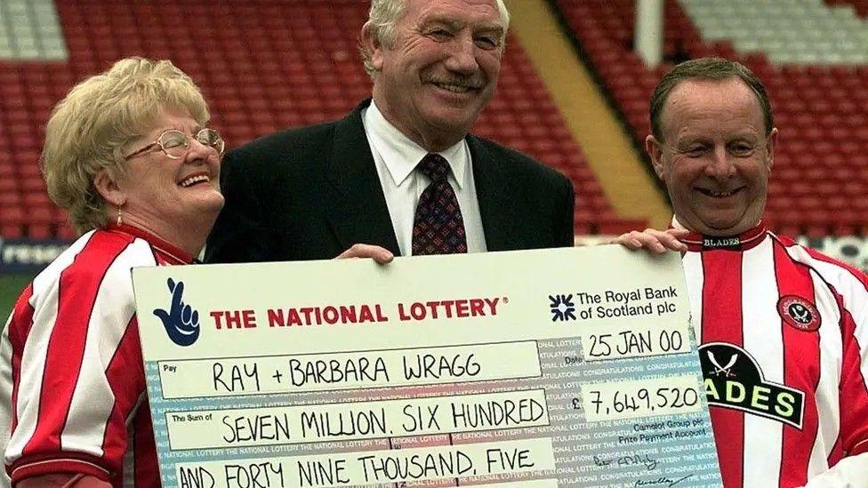 A couple dressed in red and white Sheffield United shirts stand either side of a suited man holding a giant cheque. The cheque reads: "Ray + Barbara Wragg; Seven million, six hundred and forty nine thousand' and is dated 25 January 2000.