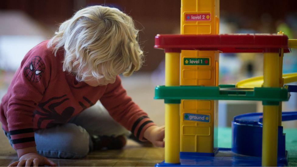 A blond-haired child, sitting on the floor, plays with a toy multi-story cark park.
