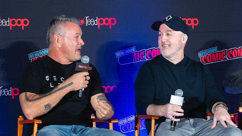 Two men, one wearing a black t-shirt and the other a black sweater and black baseball cap, sit on folding director-style chairs in front of a dark blue hoarding with a repeating pattern of New York Comic Con and Reed Pop logos printed on it. Each holds a microphone and the man on the left is