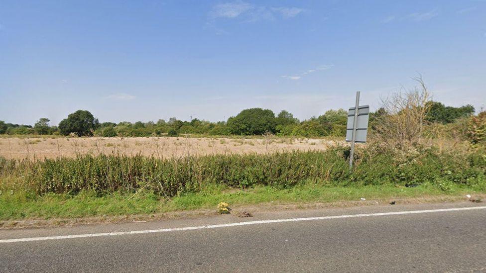 A piece of scrubby land seen from the road, which has a white line along one edge. It is edged by hedges and trees and has brown soil and weeds across the field. 