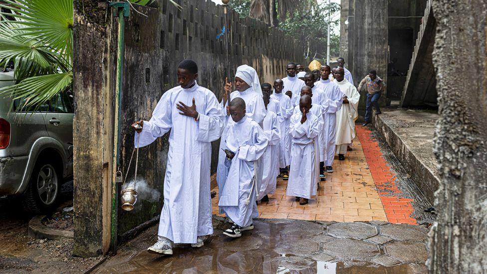 An altar boy dressed in white swings a thurible followed by other Catholic faithful and clergy also in white as they head into church at Saint Augustin of Taouyah Parish in Conakry, Guinea - Sunday 31 August 2025.