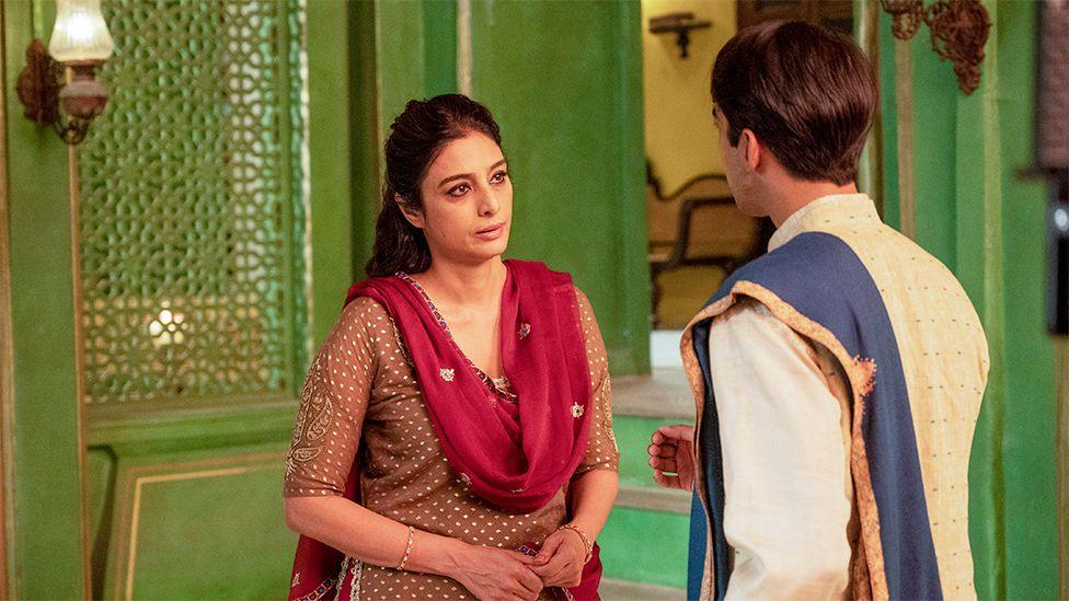 A still image of Tabu in the BBC drama A Suitable Boy. She is wearing a light brown outfit with a red shawl around her shoulders. She is standing opposite a male character, with green walls around the pair as they interact.